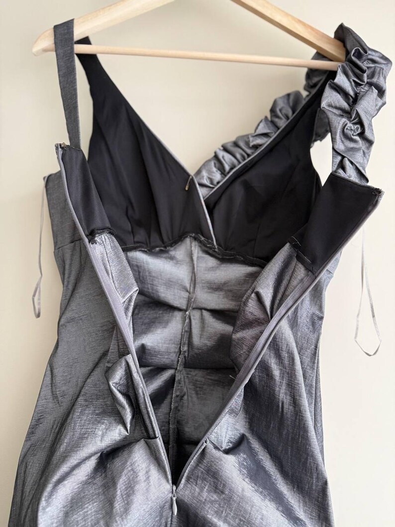 Puede incluir: Un vestido gris plateado con cuello en V y forro negro, colgado de una percha de madera. El vestido presenta una cremallera en la espalda y un detalle de volantes en el hombro. La tela tiene un brillo sutil.