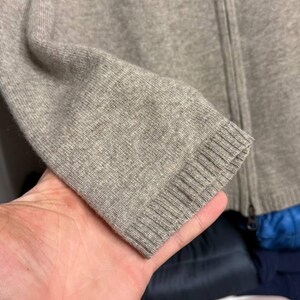 Pu&ograve; includere: Primo piano di un maglione con zip marrone chiaro. La manica &egrave; visibile, mostrando il polsino a coste e la trama del tessuto a maglia. Anche la cerniera &egrave; visibile.