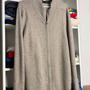 Pu&ograve; includere: Un maglione beige con zip e collo alto, appeso a una gruccia bianca. Il maglione ha una cerniera a tutta lunghezza ed &egrave; realizzato in un materiale morbido e strutturato. Il maglione &egrave; esposto contro una mensola bianca.