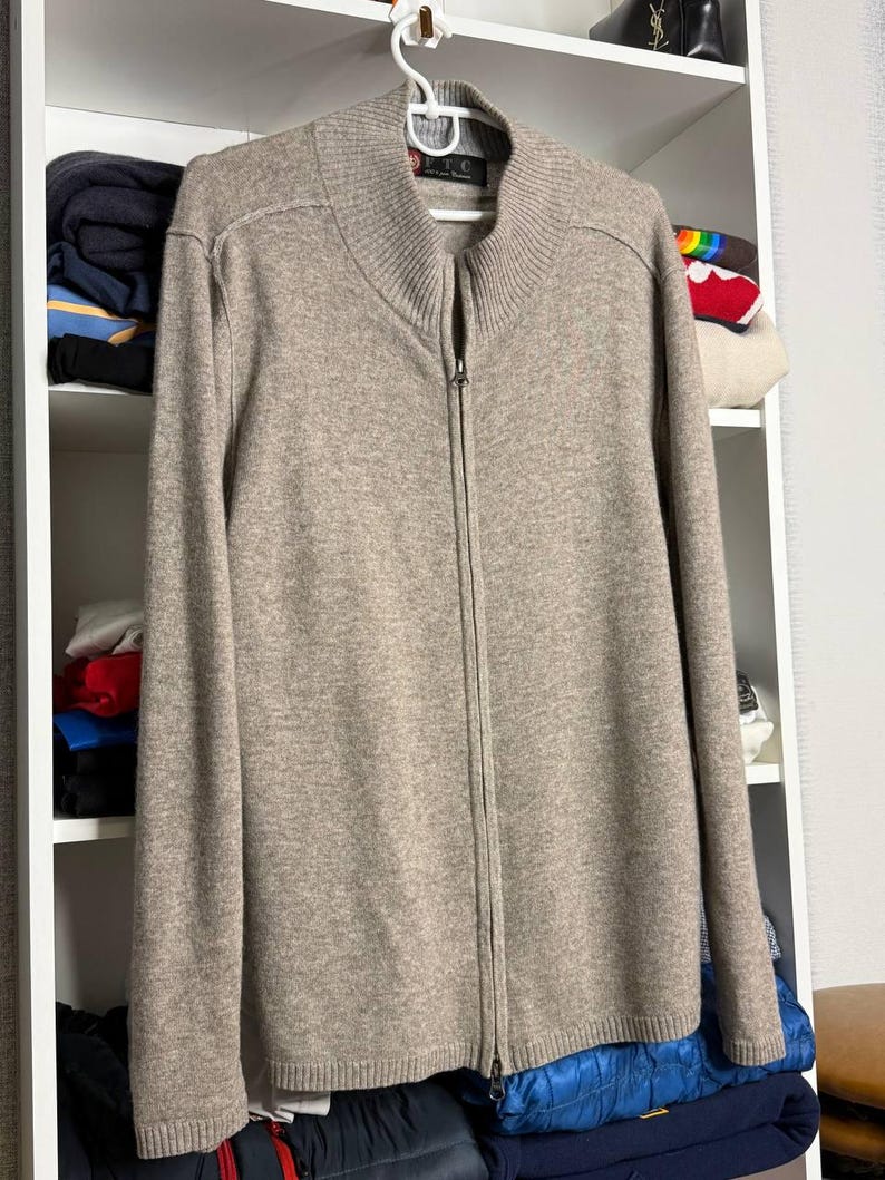 Pu&ograve; includere: Un maglione beige con zip e collo alto, appeso a una gruccia bianca. Il maglione ha una cerniera a tutta lunghezza ed &egrave; realizzato in un materiale morbido e strutturato. Il maglione &egrave; esposto contro una mensola bianca.