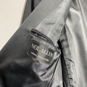 May include: Close-up of a black formal jacket interior. The lining is a shiny black fabric. A label inside the jacket reads "NEIL ALLYN FORMAL COLLECTION" in white text. The jacket appears to be made of a smooth, dark material.