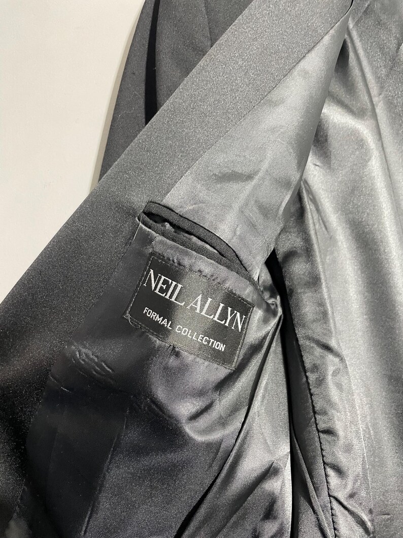 May include: Close-up of a black formal jacket interior. The lining is a shiny black fabric. A label inside the jacket reads "NEIL ALLYN FORMAL COLLECTION" in white text. The jacket appears to be made of a smooth, dark material.