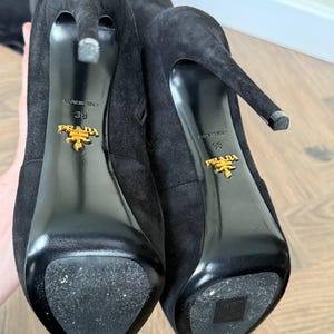 May include: Close-up of two black suede high-heeled boots. The soles are black and shiny, with the gold Prada logo and the size 38. The heels are thin and pointed. The boots are on a light brown wooden surface.