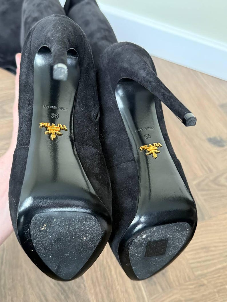 May include: Close-up of two black suede high-heeled boots. The soles are black and shiny, with the gold Prada logo and the size 38. The heels are thin and pointed. The boots are on a light brown wooden surface.