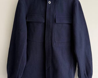 May include: A navy blue jacket with two front pockets and a collared neckline. The jacket has a button closure and long sleeves. The garment is hanging on a wooden hanger against a neutral background. The jacket appears to be made of a thick, warm material.