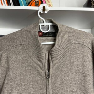 Pu&ograve; includere: Un maglione beige con zip, colletto e polsini a coste, appeso a una gruccia bianca. Il maglione ha una cerniera completa e un'etichetta visibile sul collo. Il maglione &egrave; realizzato in un materiale morbido e strutturato.