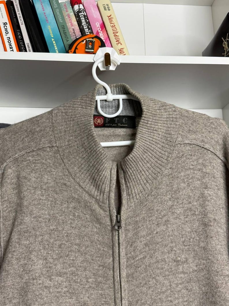Pu&ograve; includere: Un maglione beige con zip, colletto e polsini a coste, appeso a una gruccia bianca. Il maglione ha una cerniera completa e un'etichetta visibile sul collo. Il maglione &egrave; realizzato in un materiale morbido e strutturato.