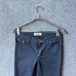 Op de afbeelding: Donkergrijze spijkerbroek met een klassiek ontwerp met vijf zakken, een metalen knoopsluiting en een wit label aan de binnenkant van de tailleband met de tekst Acne. De broek hangt aan een metalen kledinghanger met twee zilverkleurige clips.