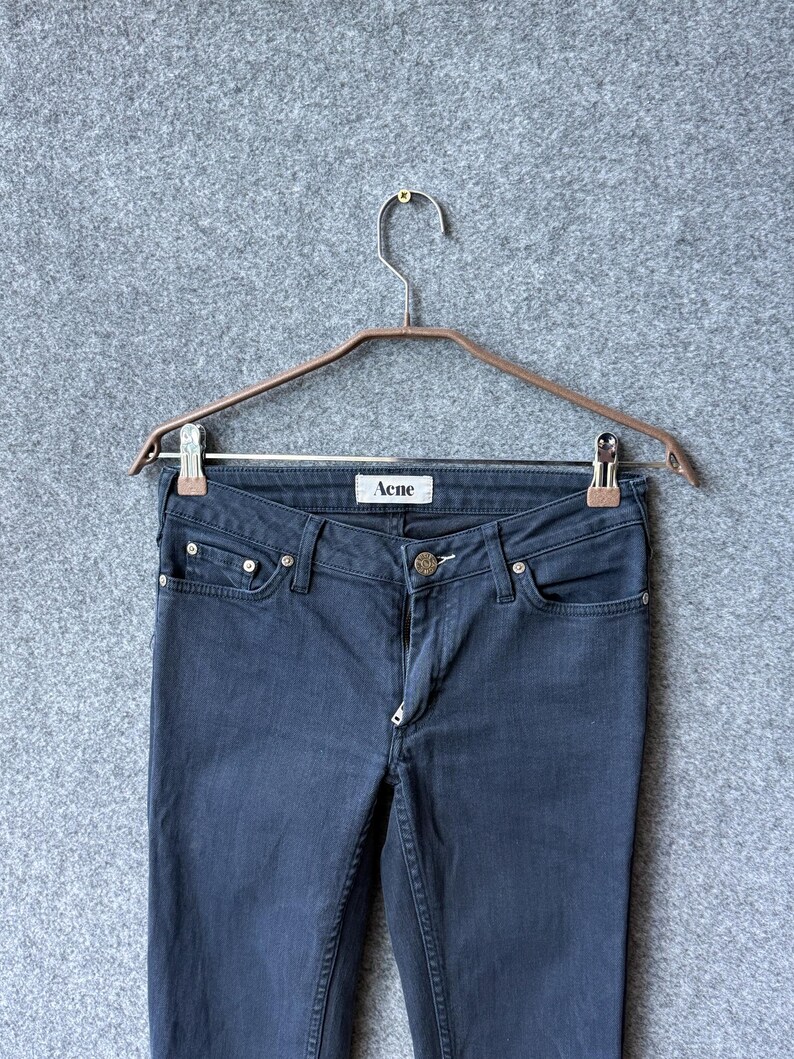 Op de afbeelding: Donkergrijze spijkerbroek met een klassiek ontwerp met vijf zakken, een metalen knoopsluiting en een wit label aan de binnenkant van de tailleband met de tekst Acne. De broek hangt aan een metalen kledinghanger met twee zilverkleurige clips.