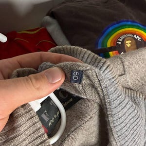 Pu&ograve; includere: Primo piano di un maglione in cashmere beige con un'etichetta taglia 50. Il maglione &egrave; appeso a una gruccia bianca. L'etichetta recita "100% puro cashmere". Altri capi di abbigliamento sono visibili sullo sfondo.
