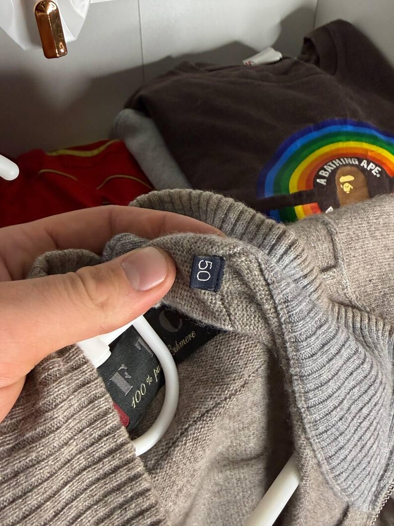 Pu&ograve; includere: Primo piano di un maglione in cashmere beige con un'etichetta taglia 50. Il maglione &egrave; appeso a una gruccia bianca. L'etichetta recita "100% puro cashmere". Altri capi di abbigliamento sono visibili sullo sfondo.