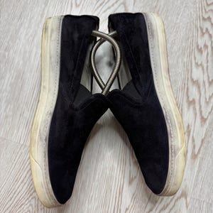 May include: Pair of black suede slip-on sneakers with off-white soles. The shoes have a worn appearance, with visible wear on the soles and sides. A metal shoe tree is inserted between the shoes.