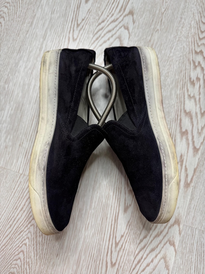 May include: Pair of black suede slip-on sneakers with off-white soles. The shoes have a worn appearance, with visible wear on the soles and sides. A metal shoe tree is inserted between the shoes.