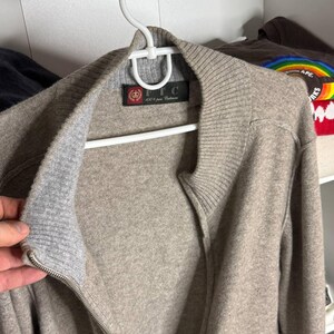 Pu&ograve; includere: Un maglione con zip marrone chiaro in 100% puro cashmere, appeso a una gruccia bianca. Il maglione ha colletto e polsini a coste e un'etichetta che recita "FTC 100% pure Cashmere".
