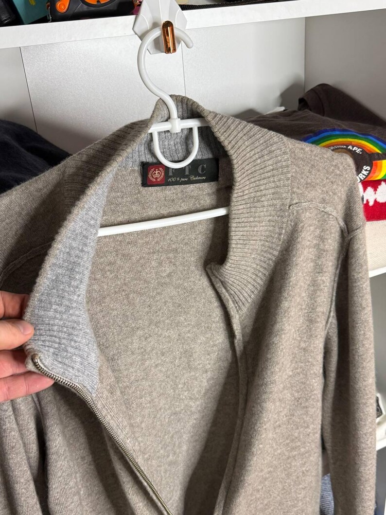 Pu&ograve; includere: Un maglione con zip marrone chiaro in 100% puro cashmere, appeso a una gruccia bianca. Il maglione ha colletto e polsini a coste e un'etichetta che recita "FTC 100% pure Cashmere".