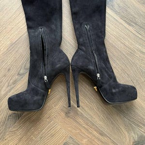 May include: A pair of black suede over-the-knee boots with stiletto heels. The boots have a side zipper and a platform sole. The heels are approximately 5 inches (12.7 cm) high. The boots are displayed on a wooden floor.