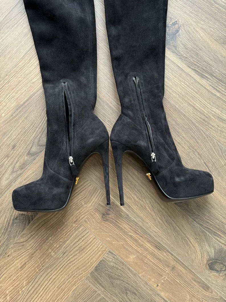 May include: A pair of black suede over-the-knee boots with stiletto heels. The boots have a side zipper and a platform sole. The heels are approximately 5 inches (12.7 cm) high. The boots are displayed on a wooden floor.