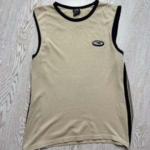 Può includere: Canotta beige con finiture nere su scollatura e giromanica. Presenta un piccolo logo sul petto e uno più grande sul retro. Strisce nere corrono lungo i lati della canotta.