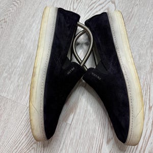 May include: A pair of black suede slip-on sneakers with white soles. The shoes have the brand name "PRADA" on the side. The soles show signs of wear. A metal shoe tree is between the shoes.