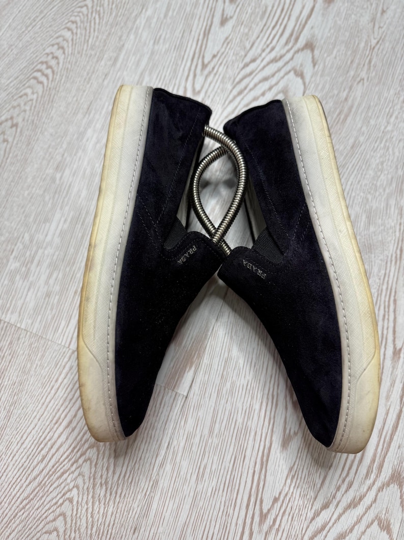 May include: A pair of black suede slip-on sneakers with white soles. The shoes have the brand name "PRADA" on the side. The soles show signs of wear. A metal shoe tree is between the shoes.