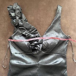 Puede incluir: Un vestido formal gris plateado con cuello en V y un solo tirante con volantes. El vestido presenta un adorno en forma de rosa en el busto y un corpi&ntilde;o ajustado. Una cinta m&eacute;trica se coloca sobre el busto.
