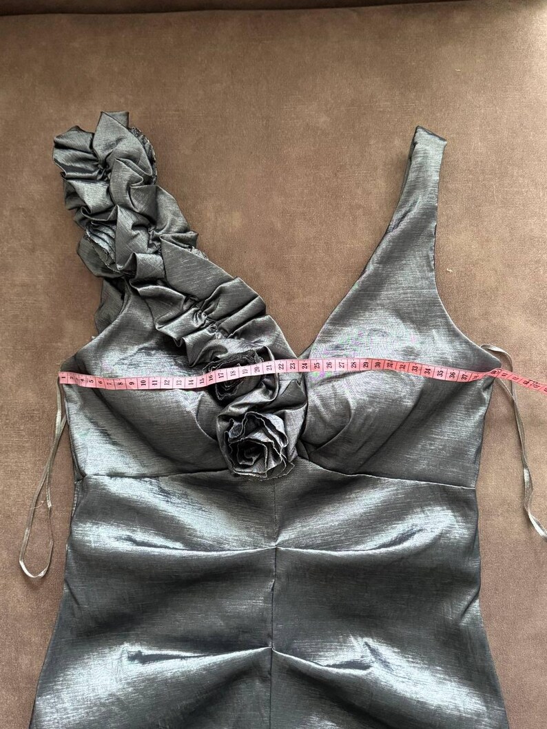 Puede incluir: Un vestido formal gris plateado con cuello en V y un solo tirante con volantes. El vestido presenta un adorno en forma de rosa en el busto y un corpi&ntilde;o ajustado. Una cinta m&eacute;trica se coloca sobre el busto.