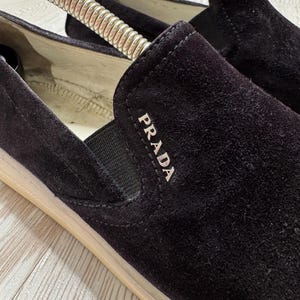 May include: Close-up of black suede slip-on shoes with the brand name "PRADA" in white lettering. The shoes have a beige sole and a black elastic band. The shoes are on a light-colored surface.