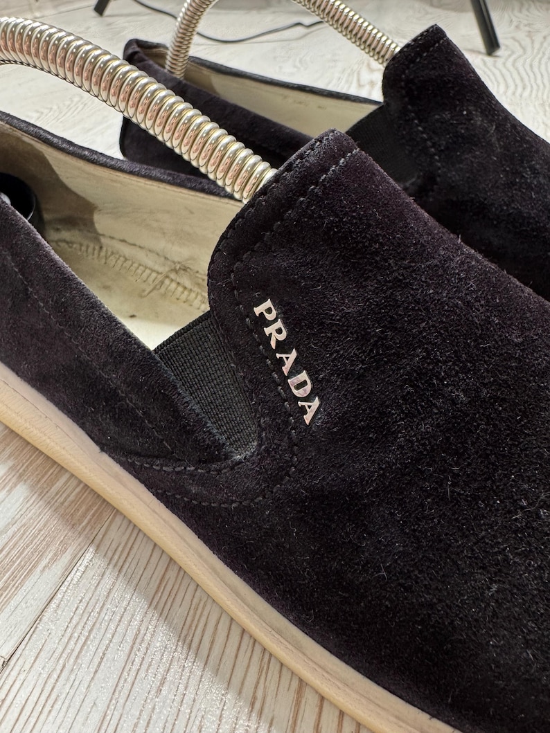 May include: Close-up of black suede slip-on shoes with the brand name "PRADA" in white lettering. The shoes have a beige sole and a black elastic band. The shoes are on a light-colored surface.