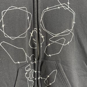 Pode incluir: Casaco preto com z&iacute;per e um design de caveira abstrata branca. O gr&aacute;fico apresenta formas e linhas geom&eacute;tricas, criando uma est&eacute;tica &uacute;nica e ousada. O casaco tem um bolso frontal e um z&iacute;per completo.