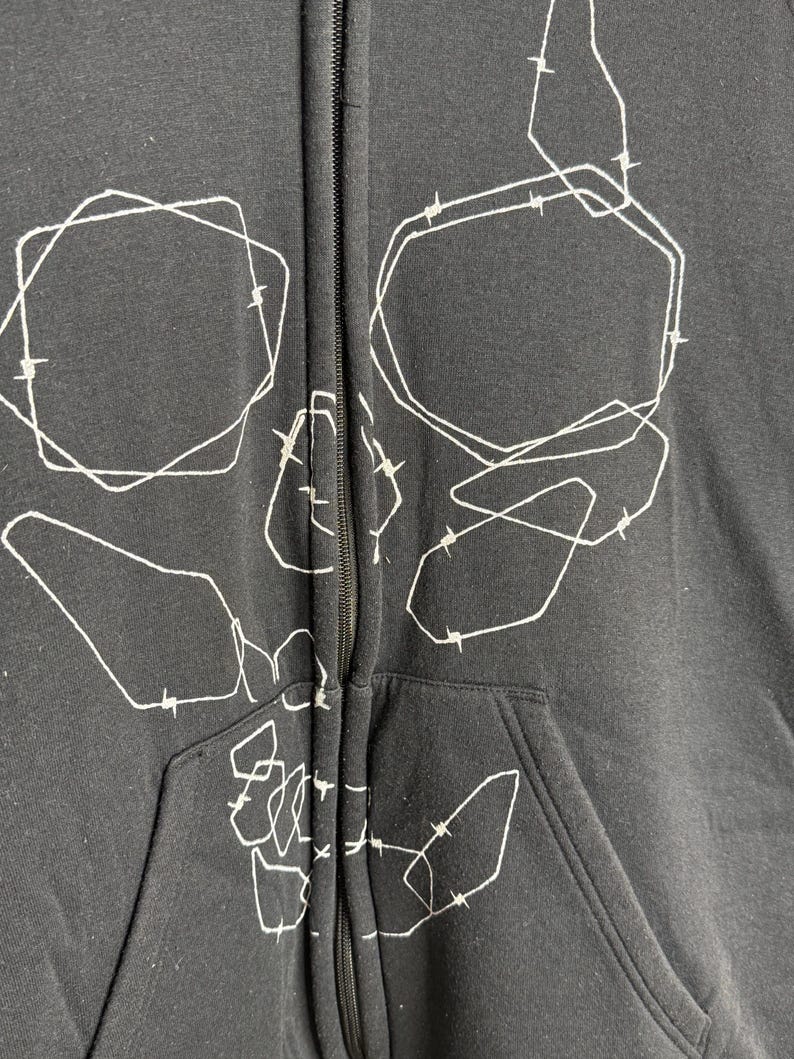 Pode incluir: Casaco preto com z&iacute;per e um design de caveira abstrata branca. O gr&aacute;fico apresenta formas e linhas geom&eacute;tricas, criando uma est&eacute;tica &uacute;nica e ousada. O casaco tem um bolso frontal e um z&iacute;per completo.