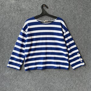 May include: A long-sleeve shirt featuring a horizontal blue and white striped pattern. The garment has a round neckline and three metallic buttons positioned vertically along the cuff of the right sleeve, hanging on a black plastic hanger.