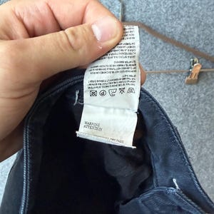Op de afbeelding: Een donkerblauw kledingstuk met een wit waslabel aan de binnenzijde met gedrukte tekst en wassymbolen. Het label bevat instructies in meerdere talen, een waarschuwingssectie en de tekst ATTENTION en WARNING onderaan het label.