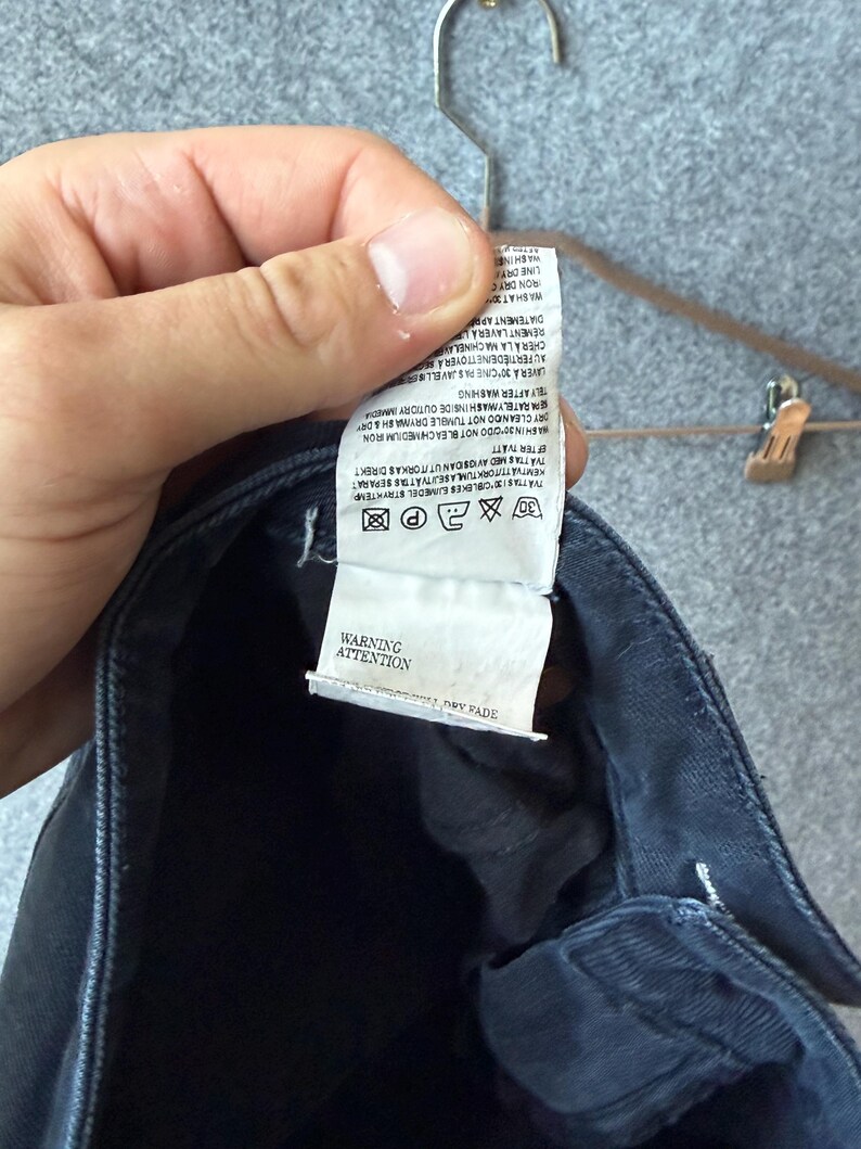 Op de afbeelding: Een donkerblauw kledingstuk met een wit waslabel aan de binnenzijde met gedrukte tekst en wassymbolen. Het label bevat instructies in meerdere talen, een waarschuwingssectie en de tekst ATTENTION en WARNING onderaan het label.