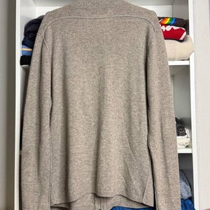 Pu&ograve; includere: Un maglione beige con zip &egrave; appeso a una gruccia bianca. Il maglione ha un collo alto e una cerniera a tutta lunghezza. Il materiale sembra essere una maglia morbida. Il maglione &egrave; esposto contro una mensola bianca con vari oggetti.