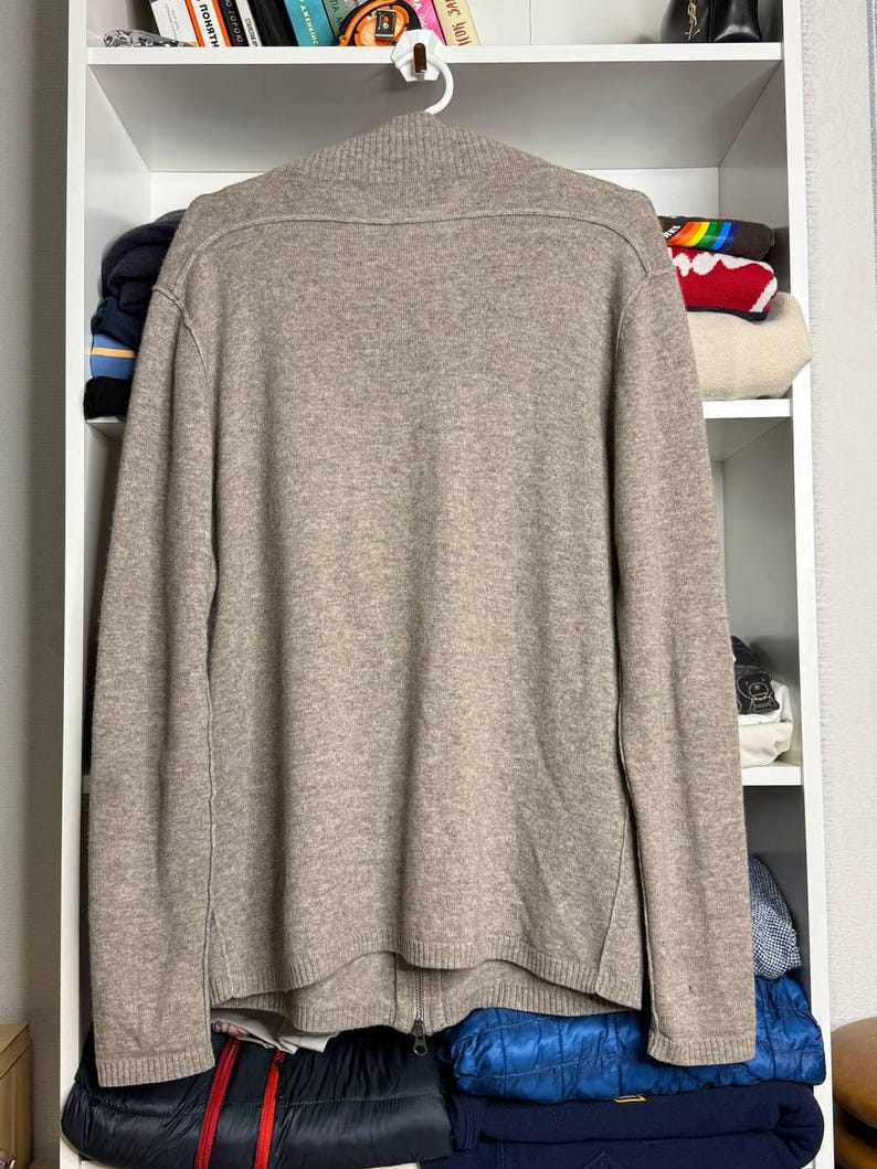 Pu&ograve; includere: Un maglione beige con zip &egrave; appeso a una gruccia bianca. Il maglione ha un collo alto e una cerniera a tutta lunghezza. Il materiale sembra essere una maglia morbida. Il maglione &egrave; esposto contro una mensola bianca con vari oggetti.