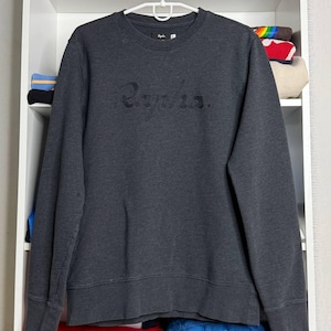 May include: Dark gray sweatshirt with the brand name "Rapha" embroidered across the chest. The crew neck sweatshirt is hanging on a white hanger against a white shelf. The shelf contains folded clothing and books.