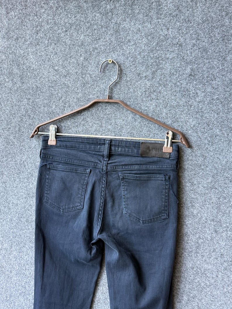 Op de afbeelding: Een donkergrijze spijkerbroek van denim met twee achterzakken, een tailleband met riemlussen en een rechthoekige leren patch aan de rechterachterkant met de merknaam Acne Studios in een subtiel, donker lettertype gedrukt.
