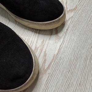 May include: Close-up of two black suede slip-on sneakers with off-white soles. The shoes are set against a light wood-grain background, showcasing the texture of the suede and the worn appearance of the soles.