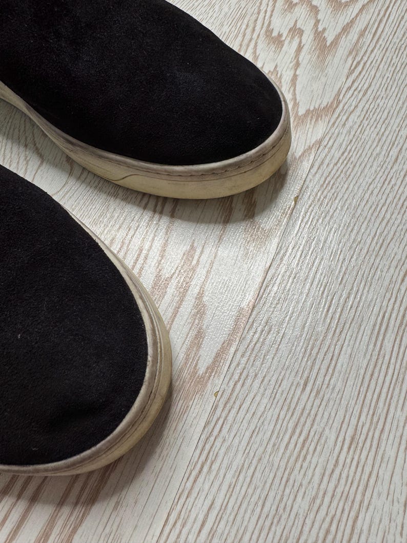 May include: Close-up of two black suede slip-on sneakers with off-white soles. The shoes are set against a light wood-grain background, showcasing the texture of the suede and the worn appearance of the soles.