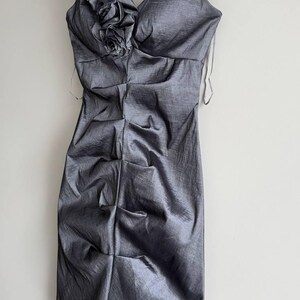 Puede incluir: Un vestido gris sin mangas con cuello en V y corpi&ntilde;o fruncido. El vestido presenta un acento floral decorativo en el hombro y cuelga de una percha de madera. La tela tiene un brillo sutil.