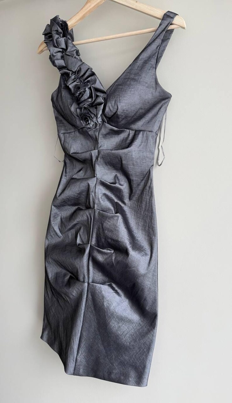 Puede incluir: Un vestido gris sin mangas con cuello en V y corpi&ntilde;o fruncido. El vestido presenta un acento floral decorativo en el hombro y cuelga de una percha de madera. La tela tiene un brillo sutil.
