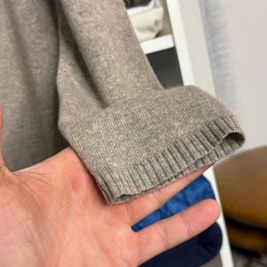 Pu&ograve; includere: Primo piano di una manica di maglione color beige. Il polsino a coste &egrave; visibile e il tessuto appare morbido e lavorato a maglia. La manica &egrave; tenuta da una mano, con uno sfondo sfocato.
