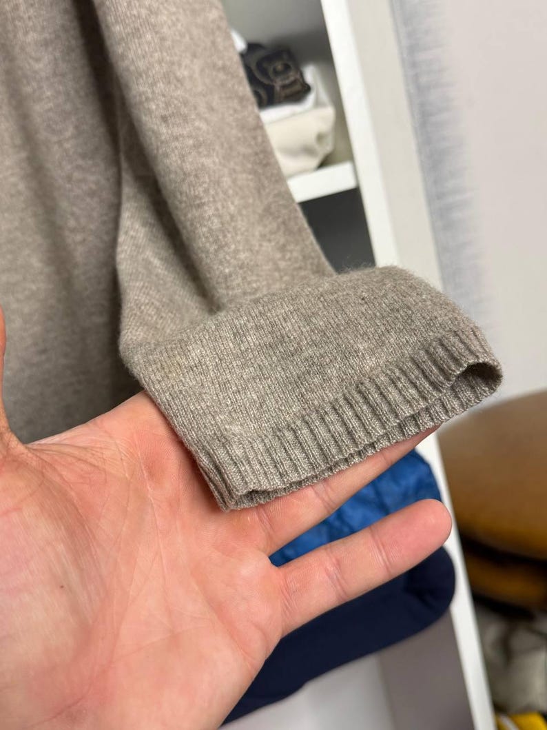 Pu&ograve; includere: Primo piano di una manica di maglione color beige. Il polsino a coste &egrave; visibile e il tessuto appare morbido e lavorato a maglia. La manica &egrave; tenuta da una mano, con uno sfondo sfocato.