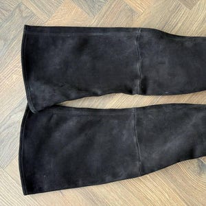 May include: Two black suede over-the-knee boots. The boots are laid flat, showcasing the full length and flared top. The suede has a soft, matte finish, and the boots appear to be unworn.