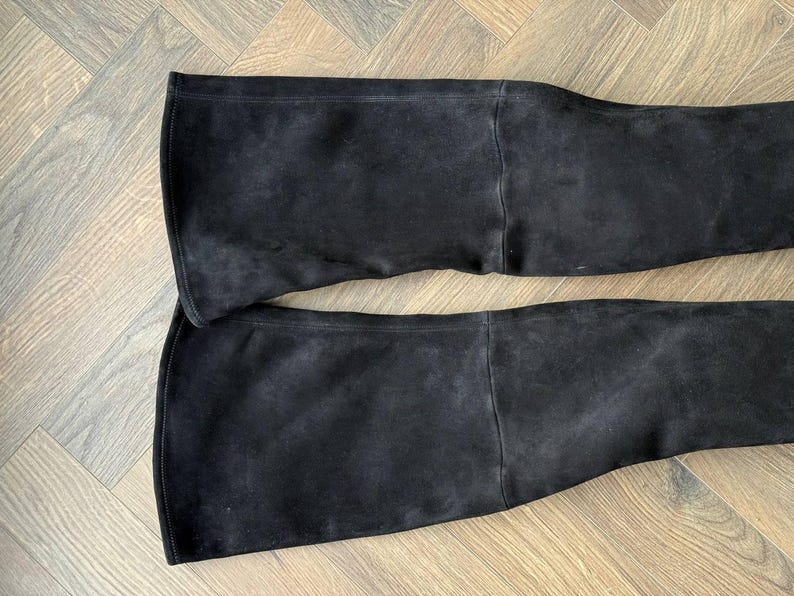 May include: Two black suede over-the-knee boots. The boots are laid flat, showcasing the full length and flared top. The suede has a soft, matte finish, and the boots appear to be unworn.