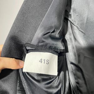 May include: Close-up of a black blazer's interior, revealing a satin lining and a label marked "41S." The fabric appears smooth and the stitching is neat. The blazer is likely part of a suit or formal wear.