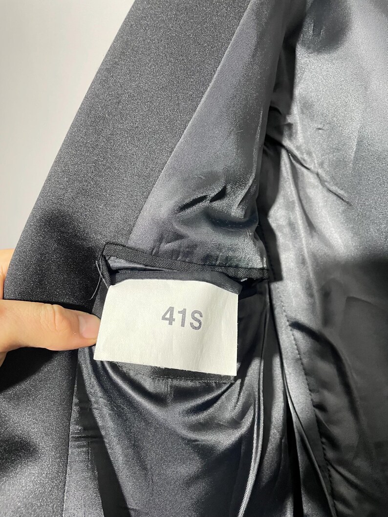 May include: Close-up of a black blazer's interior, revealing a satin lining and a label marked "41S." The fabric appears smooth and the stitching is neat. The blazer is likely part of a suit or formal wear.