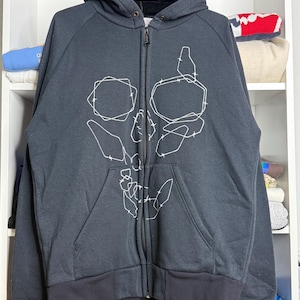 Pode incluir: Casaco preto com z&iacute;per e design de caveira em contorno branco. O casaco tem capuz, z&iacute;per completo e dois bolsos frontais. Os punhos e a bainha s&atilde;o pretos.