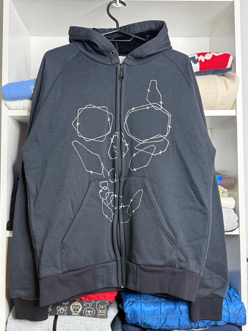 Pode incluir: Casaco preto com z&iacute;per e design de caveira em contorno branco. O casaco tem capuz, z&iacute;per completo e dois bolsos frontais. Os punhos e a bainha s&atilde;o pretos.