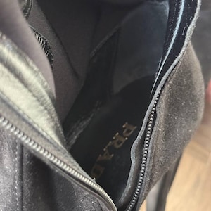 May include: Close-up of a black suede boot interior with a zipper detail. The brand name "PRADA" is visible on the insole. The boot has a high heel and a sleek, modern design.